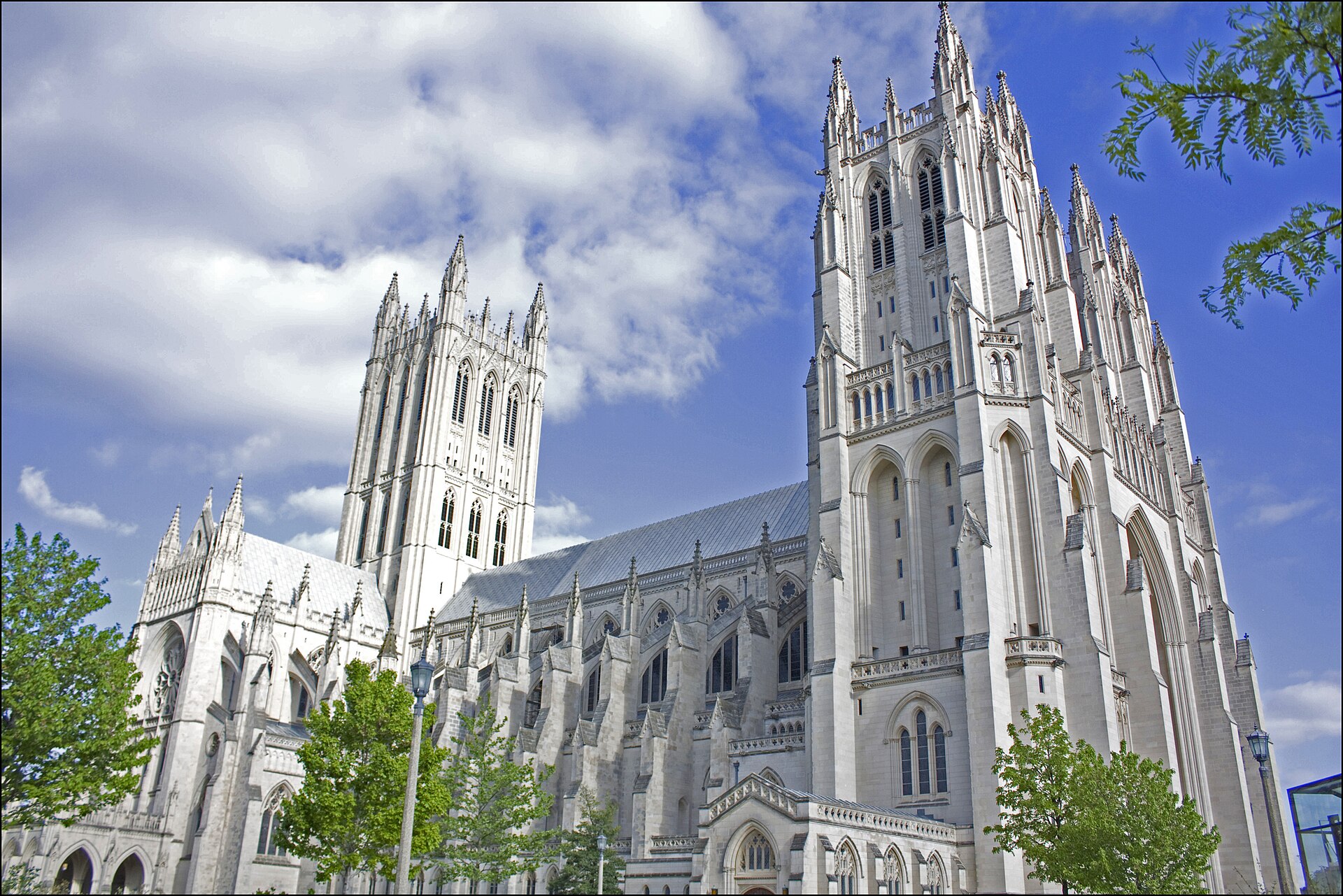Large Gothic cathedral of pale stone with a tall central tower and two west towers, seen across a lawn under a blue sky