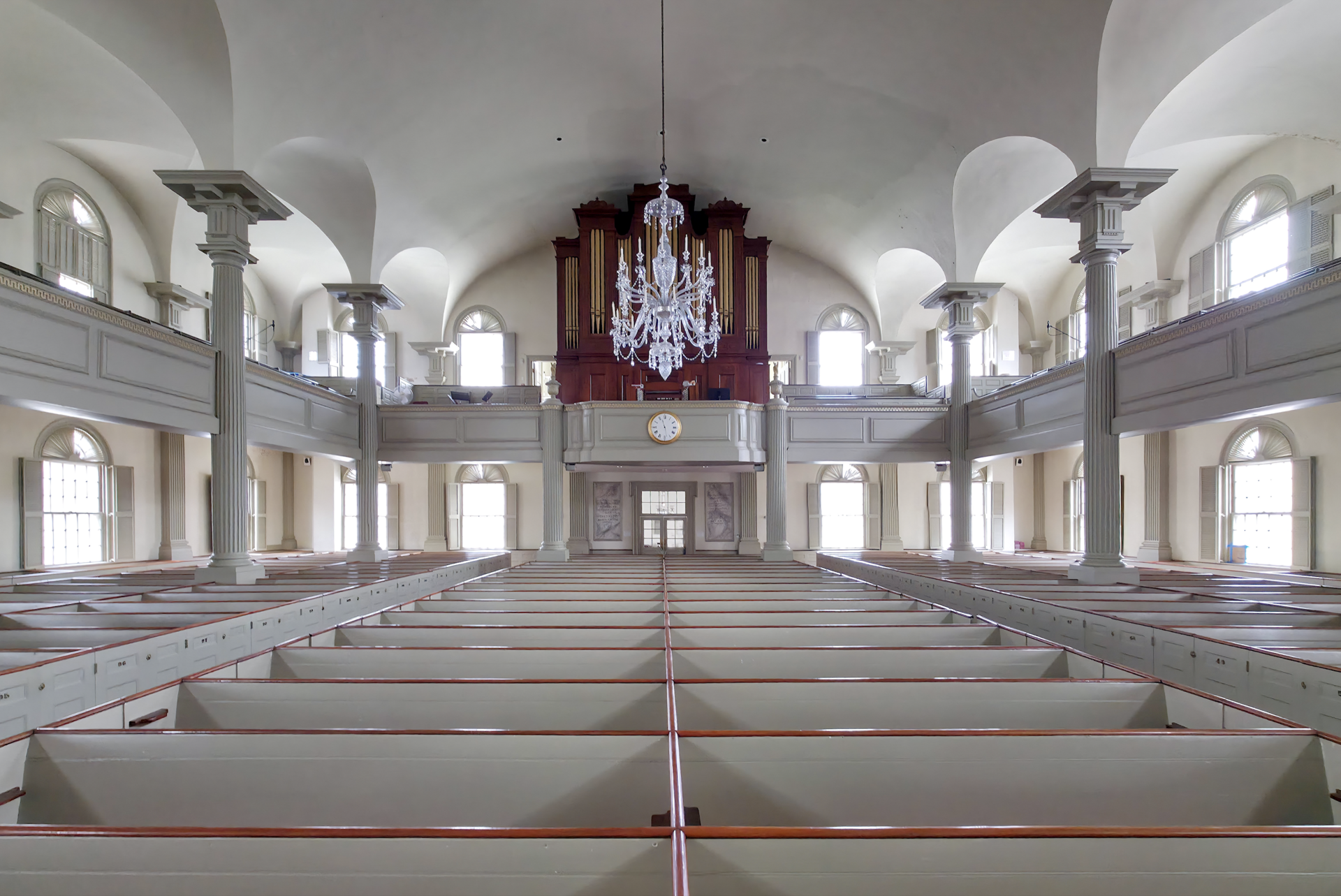 The interior of a white colonial meetinghouse with box pews, tall clear windows, a raised pulpit and a chandelier