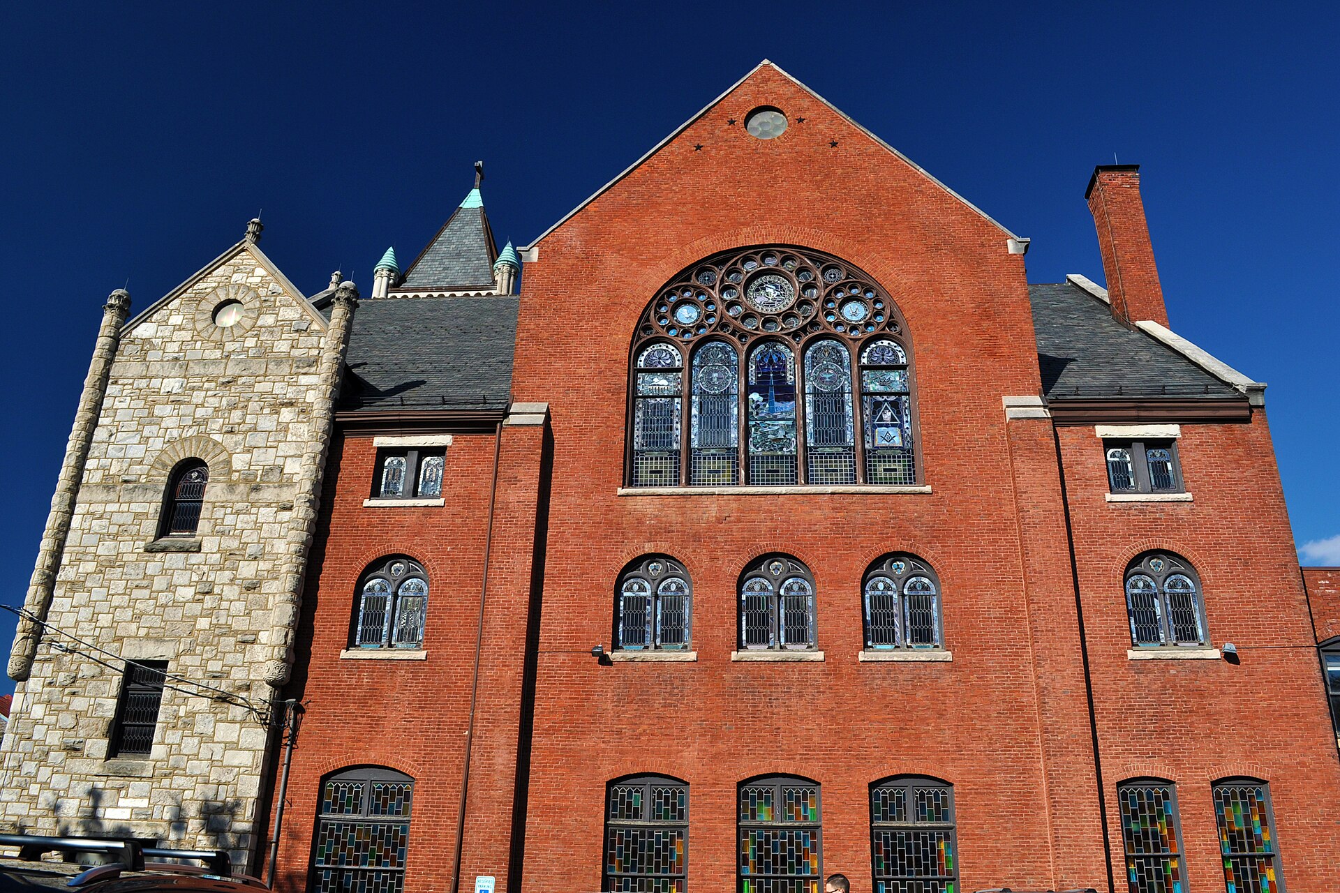 A large brick church with a tall central gable, a rose window, round-arched doors and stone steps, seen from a city sidewalk