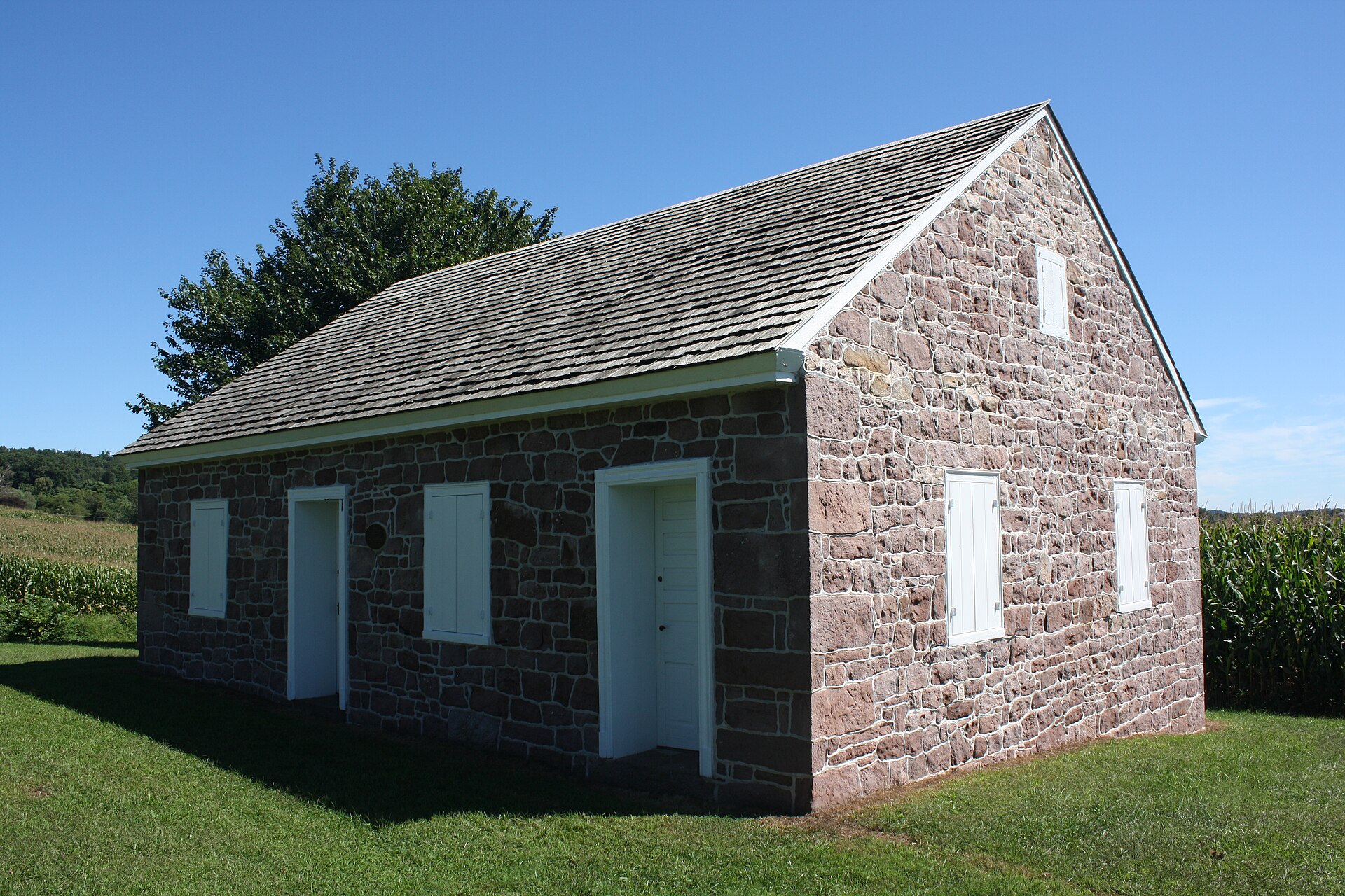 A plain rectangular brick meetinghouse with a metal roof and two front doors, standing on a lawn beside a parking area and trees