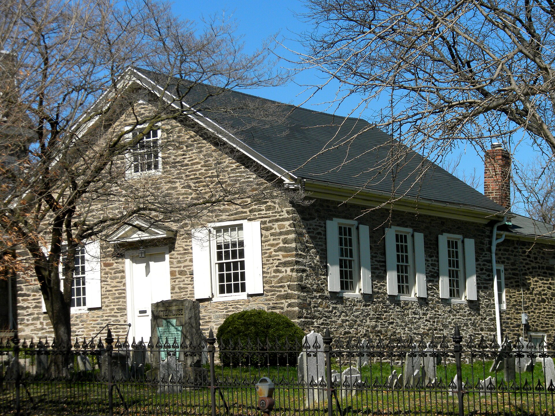 A small two-story stone building with a gabled roof and white-trimmed windows set behind a low wall on a city street