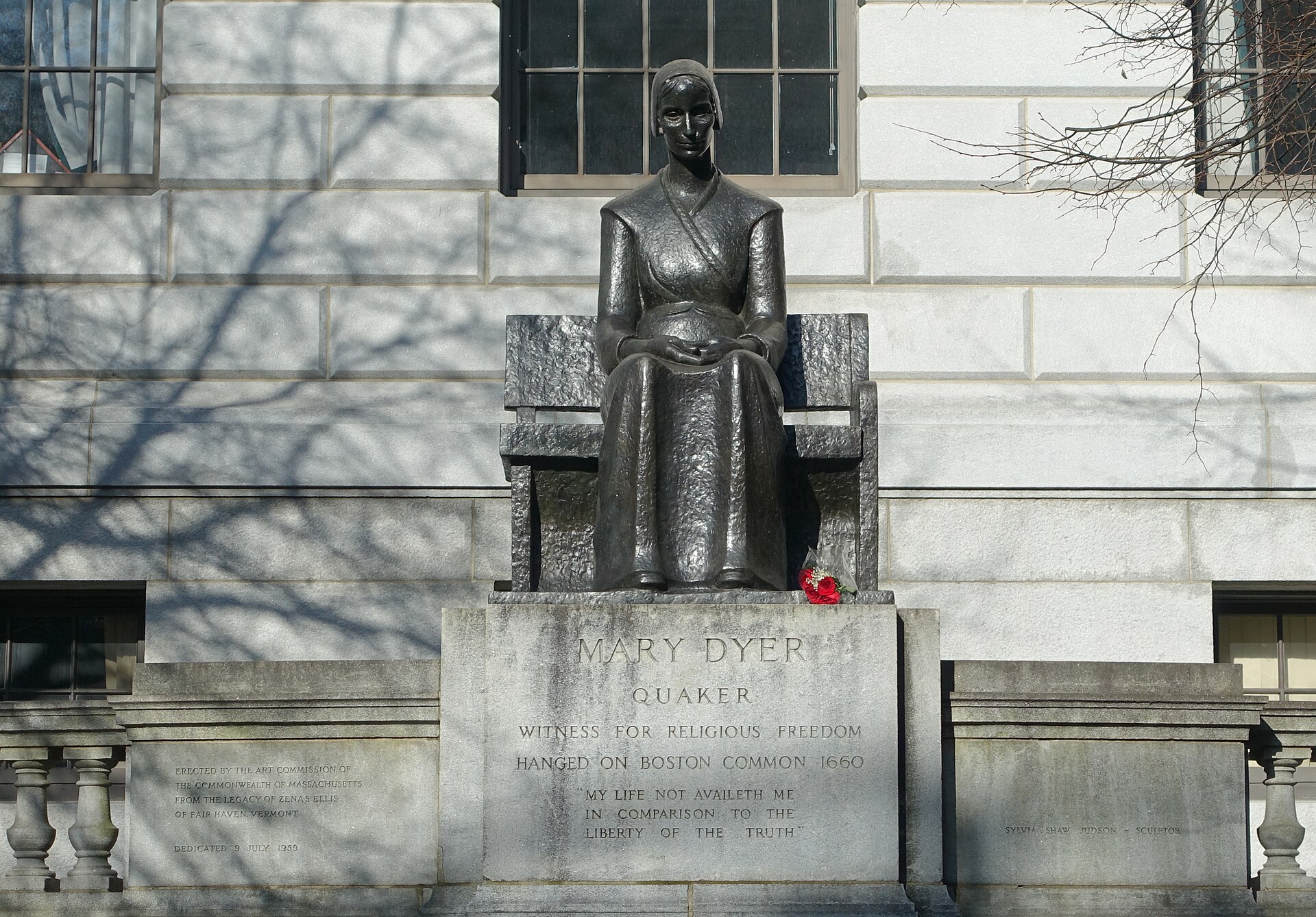 Bronze statue of a seated woman in a long plain dress and bonnet, hands folded in her lap, on a granite base in front of a stone building