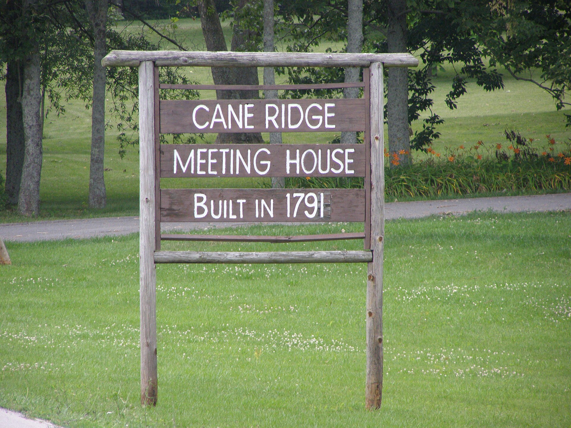 A two-story log meeting house with a wooden shingle roof, enclosed inside a larger modern stone building with tall windows