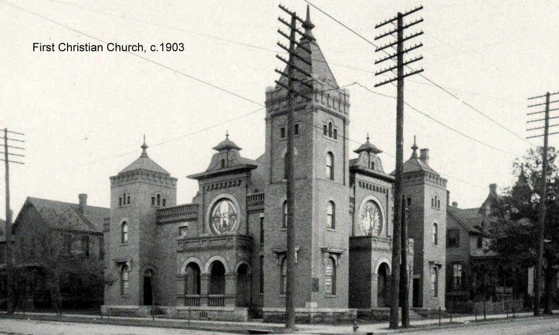 A red brick church with a squat corner bell tower, arched stained-glass windows and a small front lawn beside a road