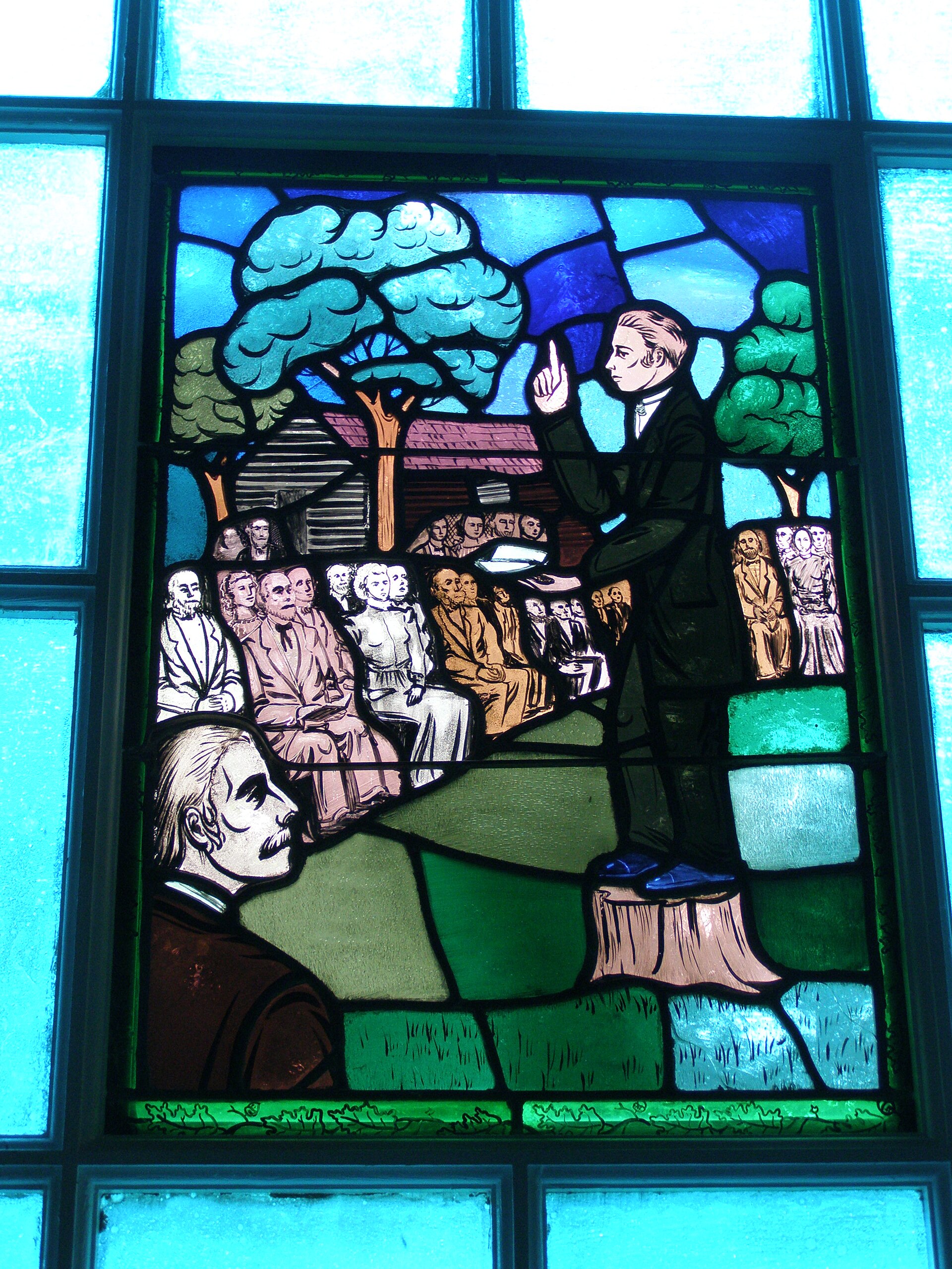A round bronze relief medallion showing a preacher standing on a tree stump with one arm raised, speaking to a crowd of people under trees