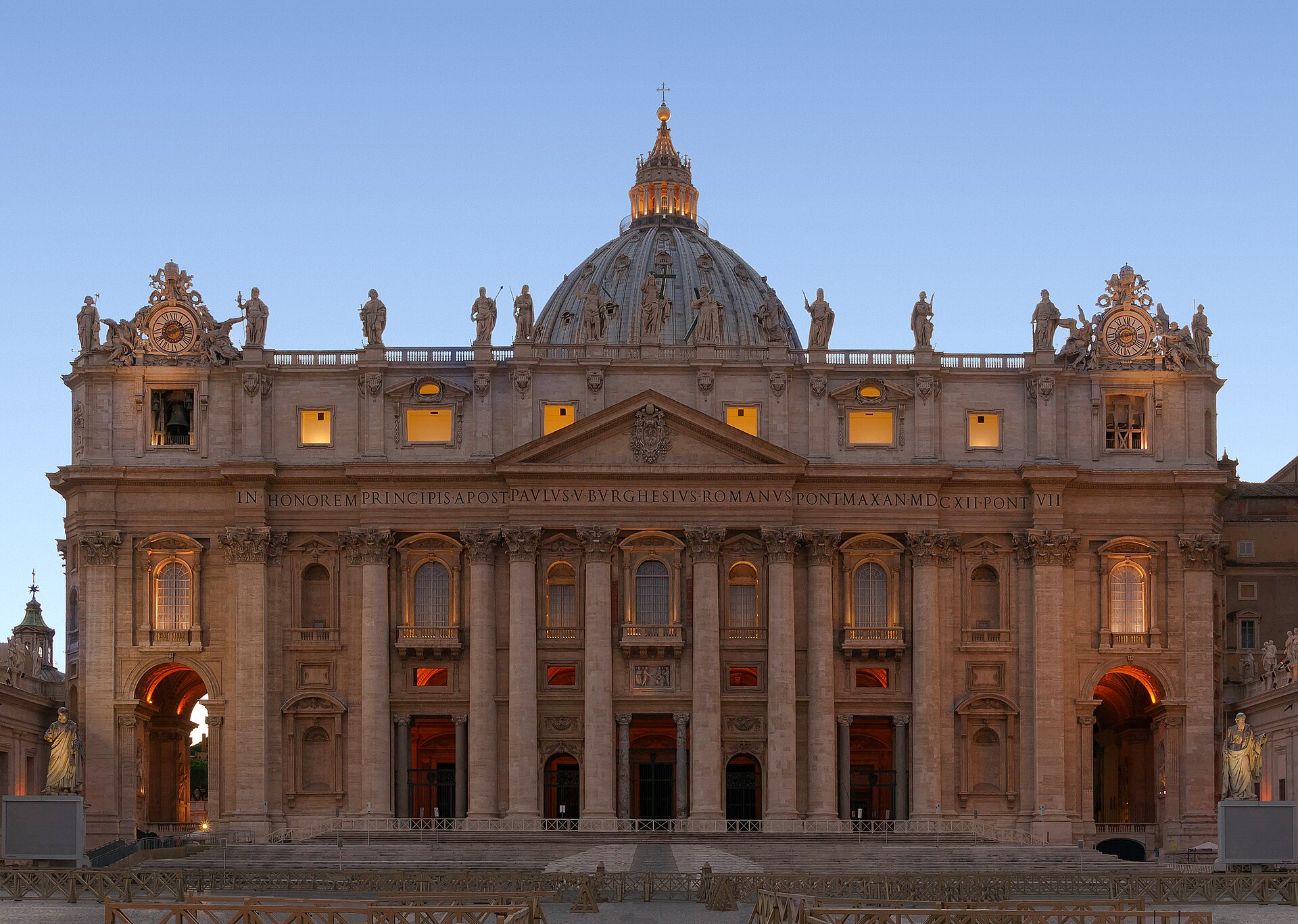 Facade of Saint Peter's Basilica in Rome at dusk, with its columned front, statues along the roofline and the great dome lit against a deep blue sky