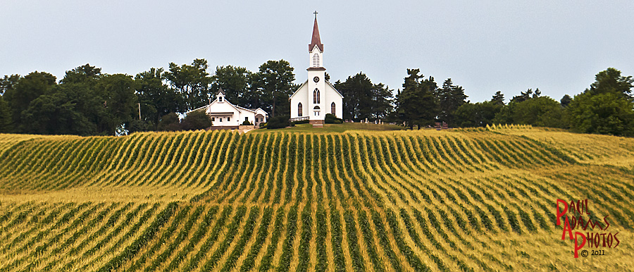 A white wooden country church with a tall steeple standing among green fields under a wide sky