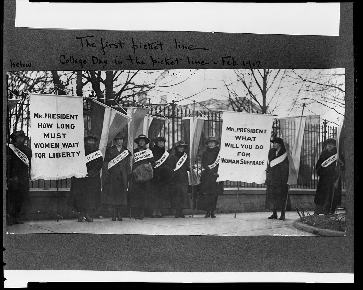Black and white 1917 photograph of women holding suffrage banners in a picket line outside the White House fence