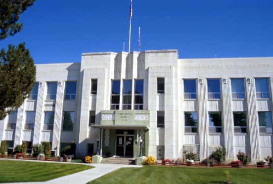 Light stone courthouse with tall vertical pilasters, a flagpole and a lawn in front