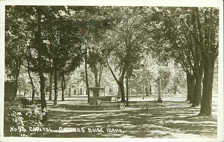 Postcard of the east lawn of the Idaho Capitol grounds in Boise around 1920, with trees and a granite monument