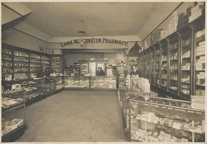 Black and white photograph of three men in white coats standing behind a wooden counter in an early twentieth century pharmacy, with shelves of bottles behind them.