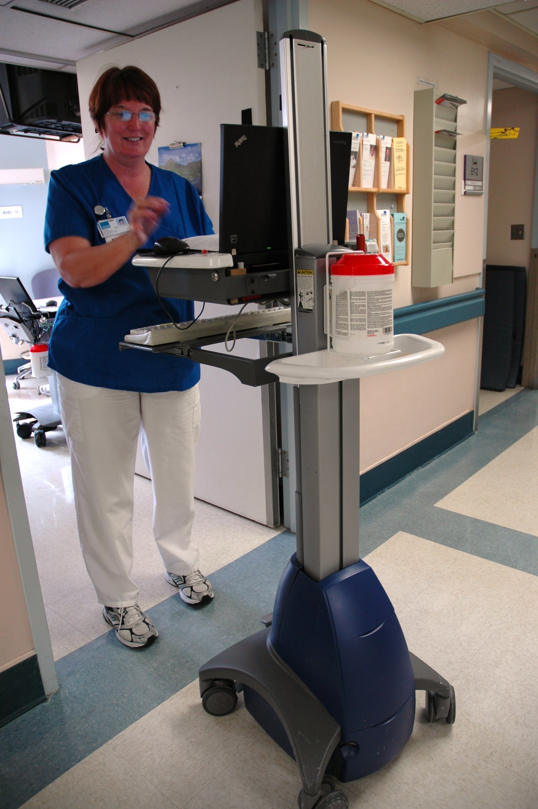 A portable computer workstation on a rolling cart in a hospital hallway, with a monitor, keyboard and a container of hand wipes, and a nurse standing nearby