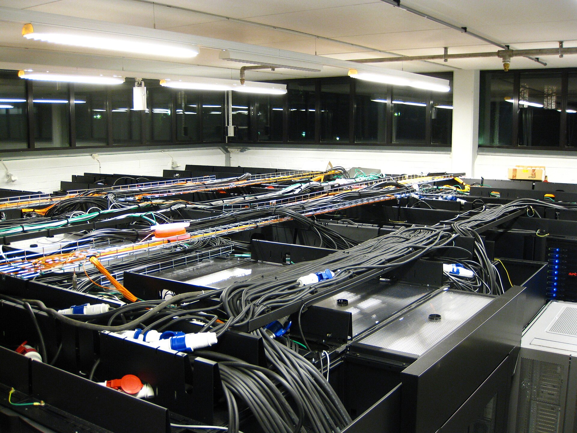 A server room with tall black computer racks, bundles of thick black power cables and orange, yellow, green and gray network cables, and pipes for a fire suppression system along the ceiling