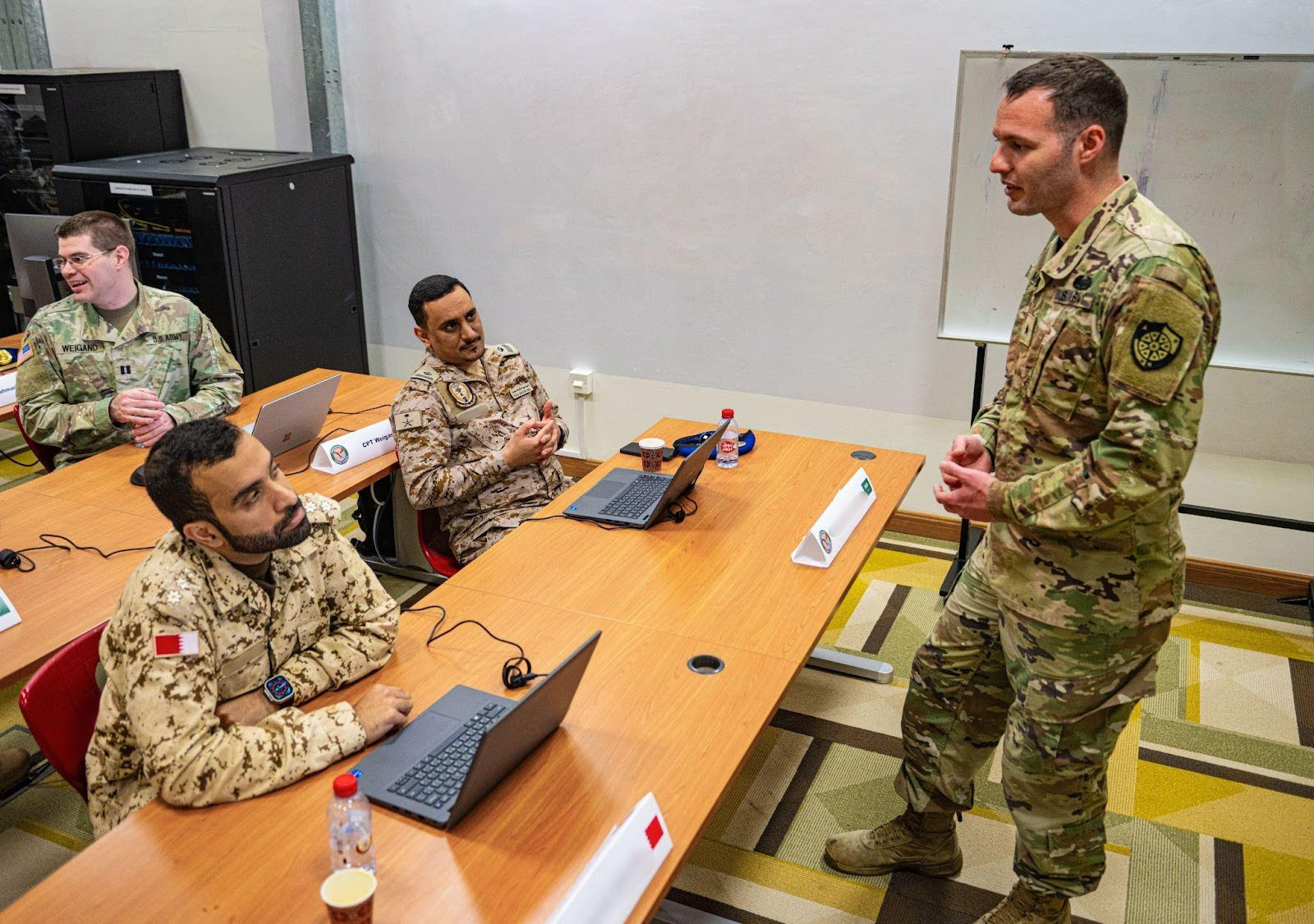 Several people in military uniforms sit around a table with laptops and papers, talking during a training workshop