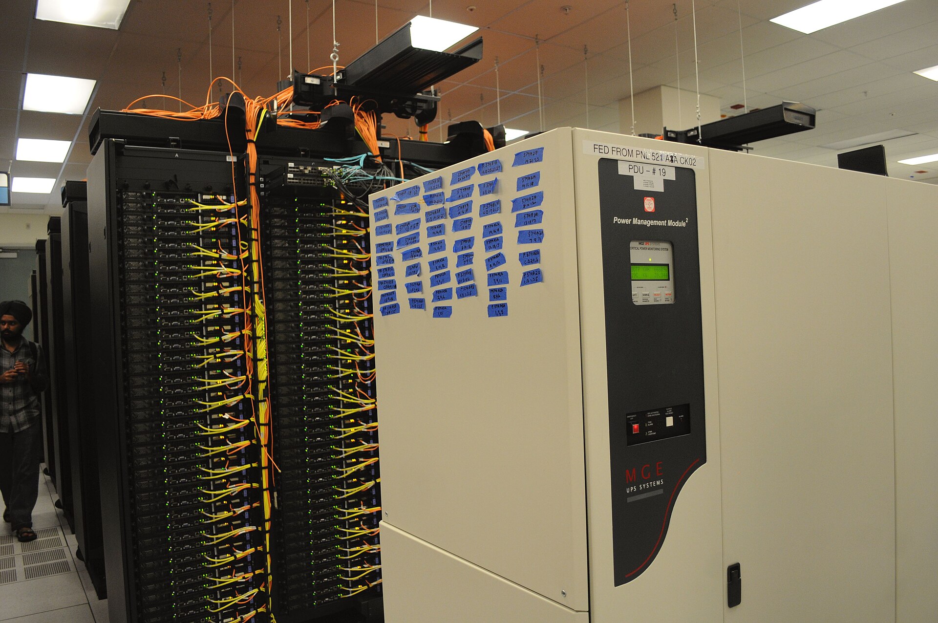 Rows of tall black server racks with network cabling and a power management module in a large data center.