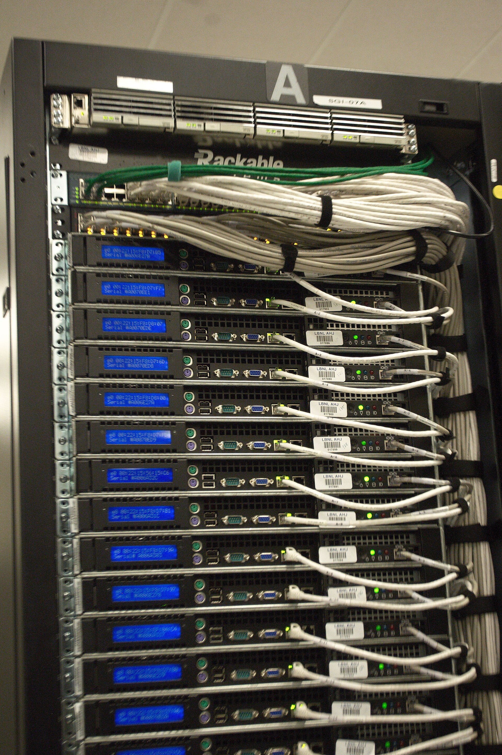 Close-up of the back of a server rack with white Ethernet cables plugged into ports and small blue LED screens showing addresses and serial numbers.