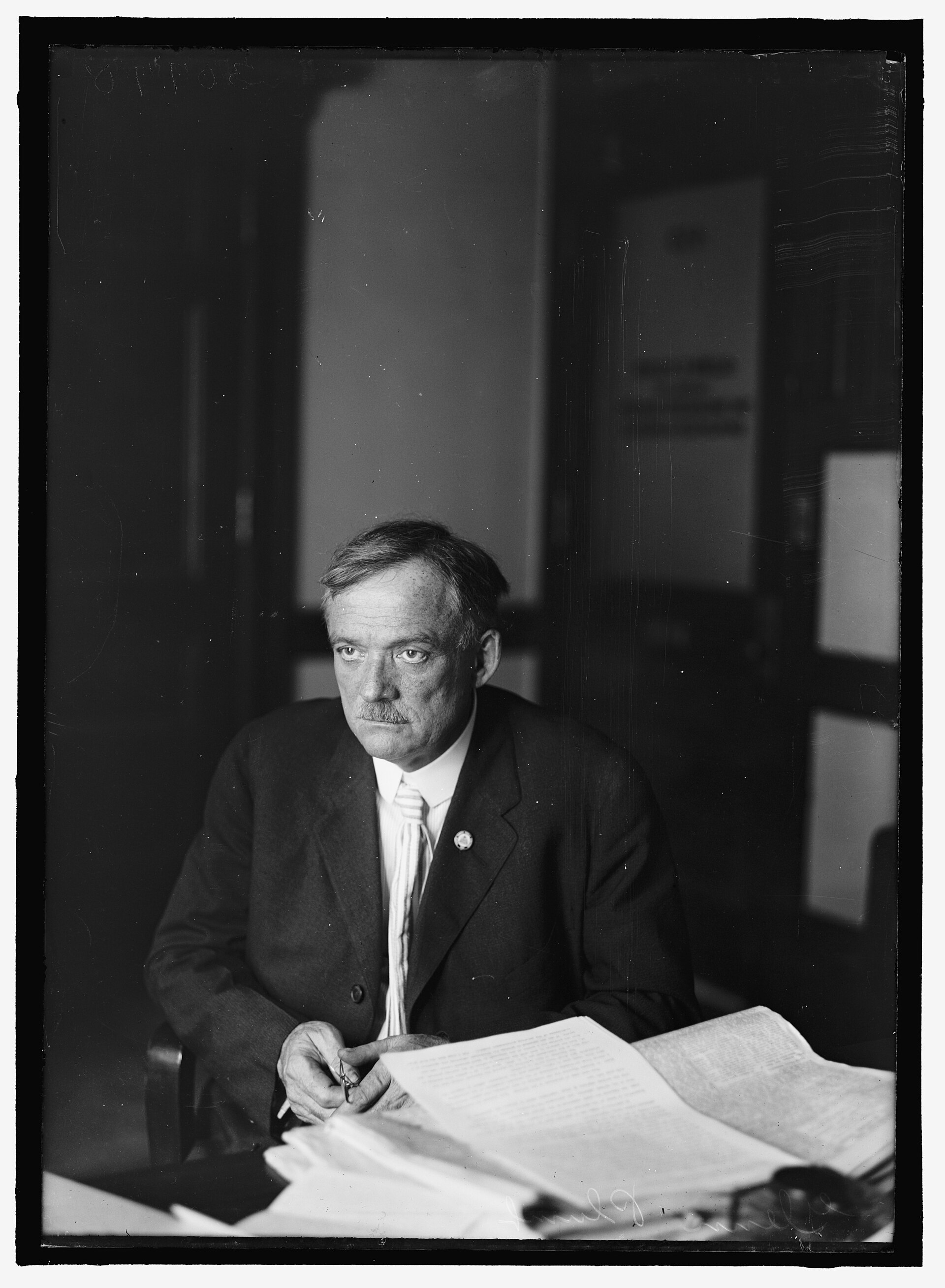 A black and white portrait of a man in a dark suit seated at a wooden office desk with papers, a telephone and books, in an early twentieth century law office.
