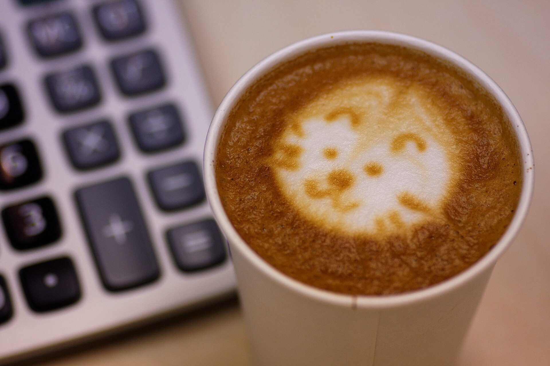 Close-up of a paper cup of latte with foam art shaped like a cat's face, beside the gray and black keys of a desk calculator.