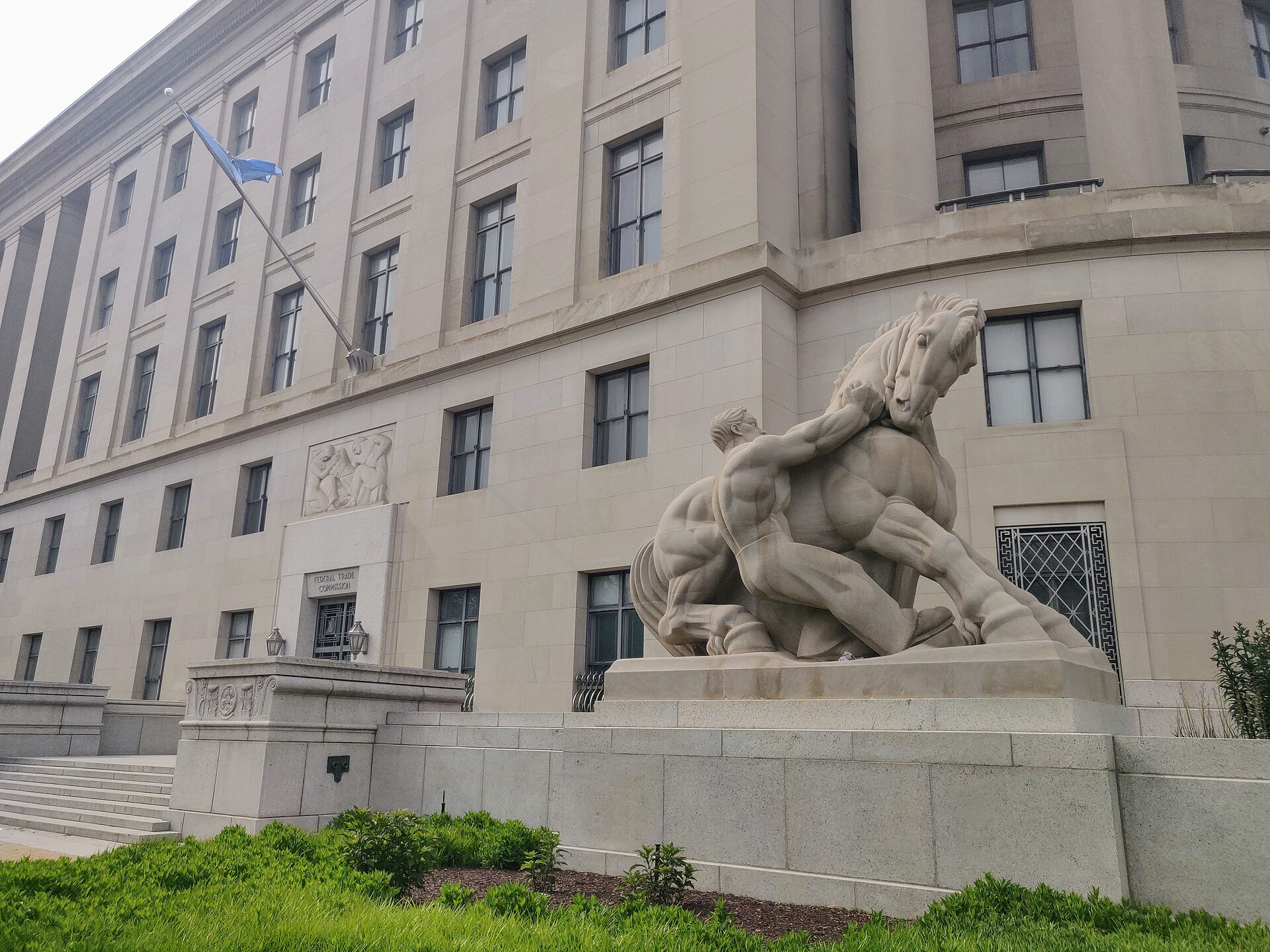 The pale stone Federal Trade Commission building in Washington, D.C., seen from the corner, with rows of tall windows, a flag on a pole and a large stone sculpture of a man holding back a rearing horse on a pedestal in front.