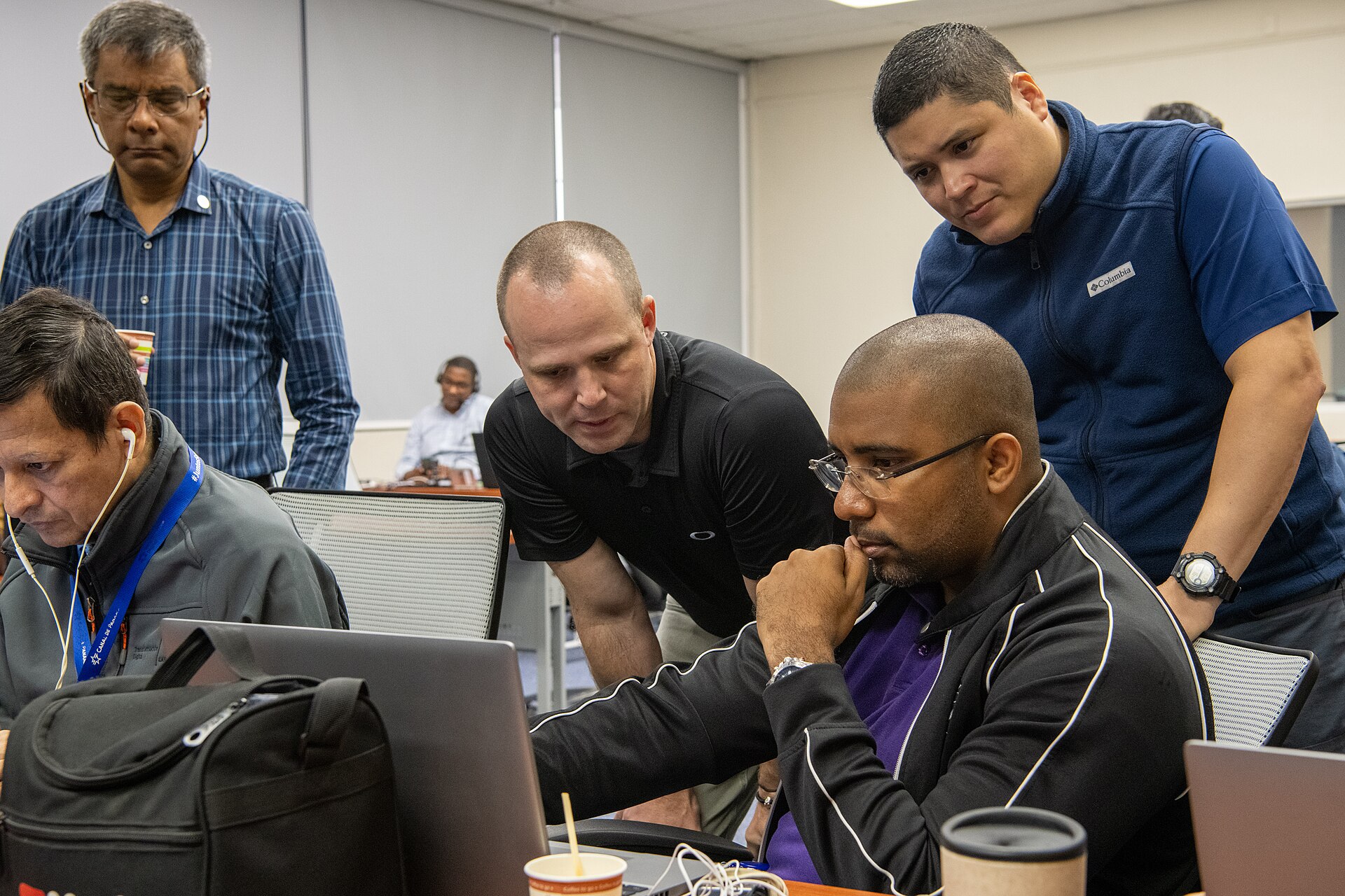 Five men gathered around laptops on a table in a conference room; one man in a black polo shirt leans over to point at a screen while the others watch, one with his hand at his chin.