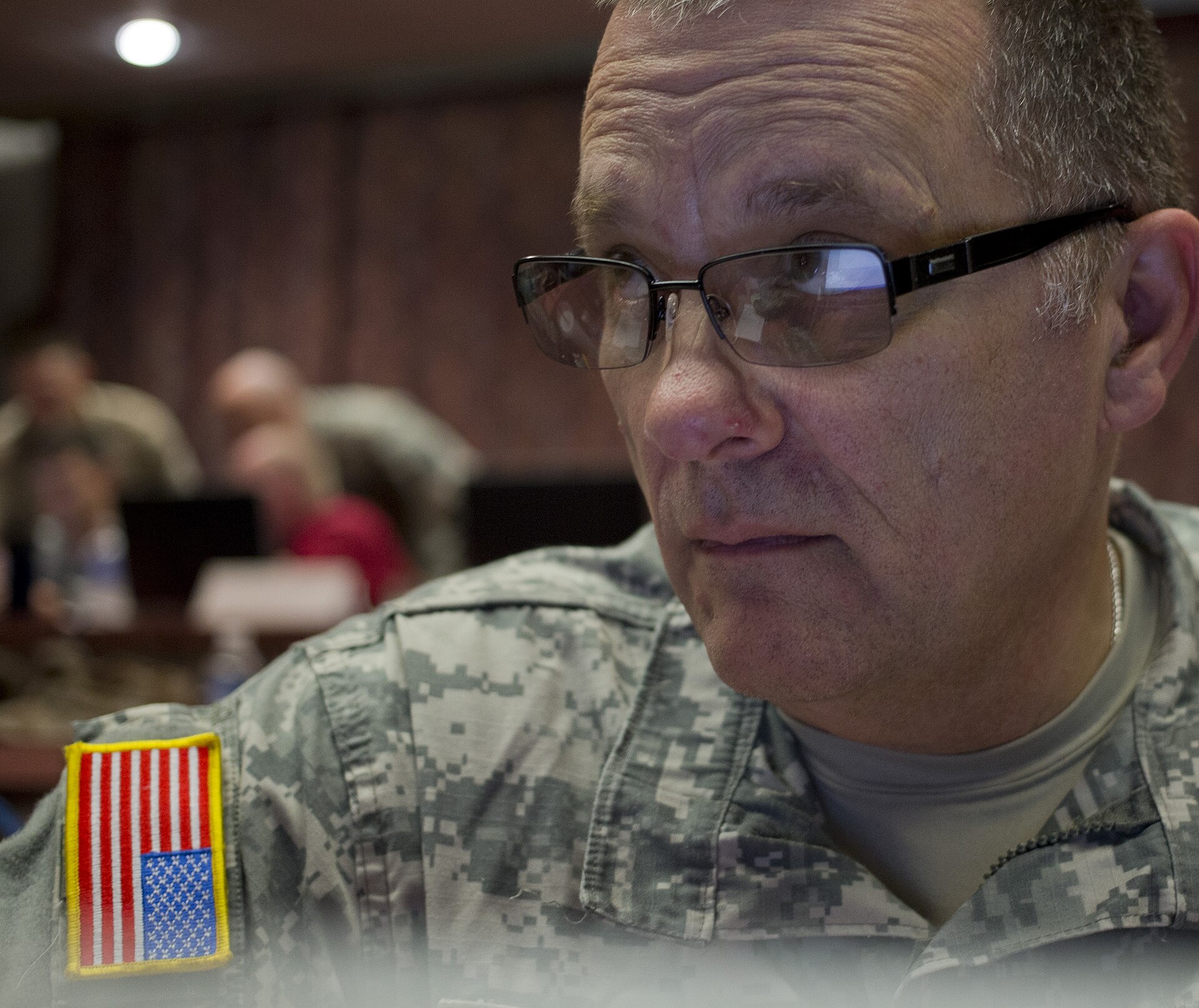 Close-up of a man in a U.S. Army camouflage uniform with an American flag patch and wire-rimmed glasses, looking at a screen, with other people working at computers in a dim room behind him.
