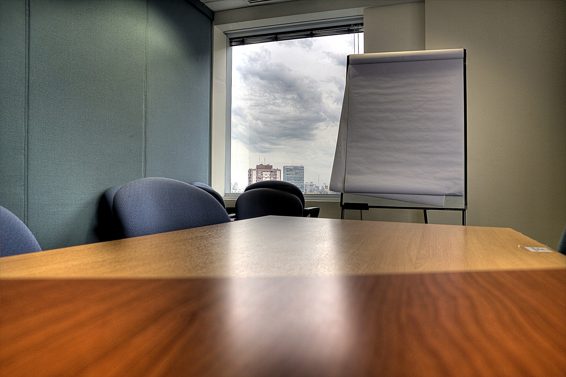 An empty conference room with a wooden table in the foreground, dark blue office chairs, a flip chart on an easel and a window looking out at city buildings under a cloudy sky.