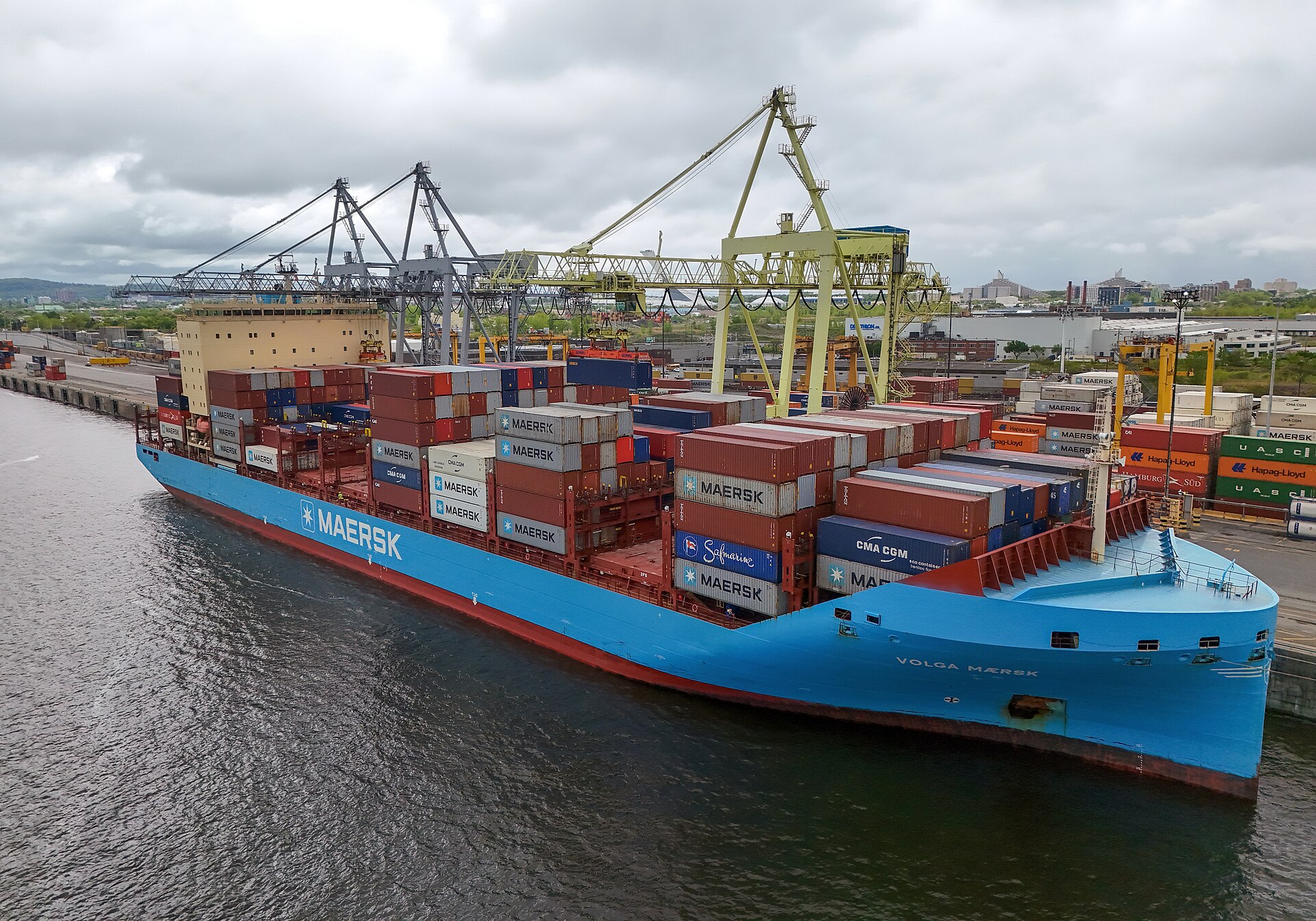 A large blue-hulled Maersk container ship loaded with stacked shipping containers, docked at a port with cranes nearby.