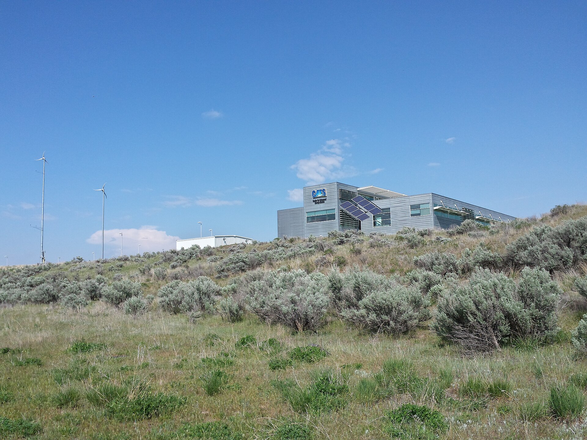 A modern gray metal-clad building with solar panels mounted on one wall sits on a low hill of sagebrush and grass under a blue sky, with two tall thin poles and a smaller white building to the left.