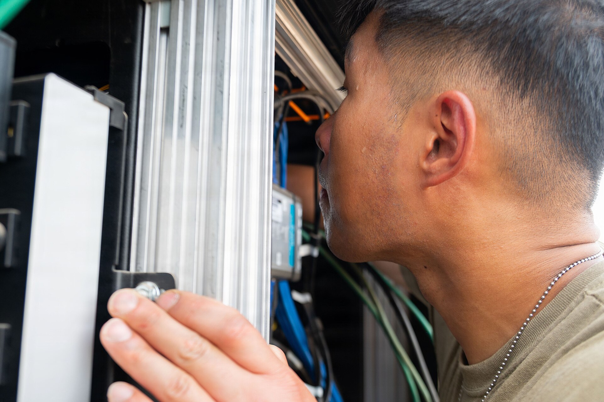 Close-up of a young man in an olive T-shirt and a chain necklace leaning his face close to an open equipment cabinet, one hand on a metal rail, with blue, green and orange cables and a small electronic module inside the cabinet.