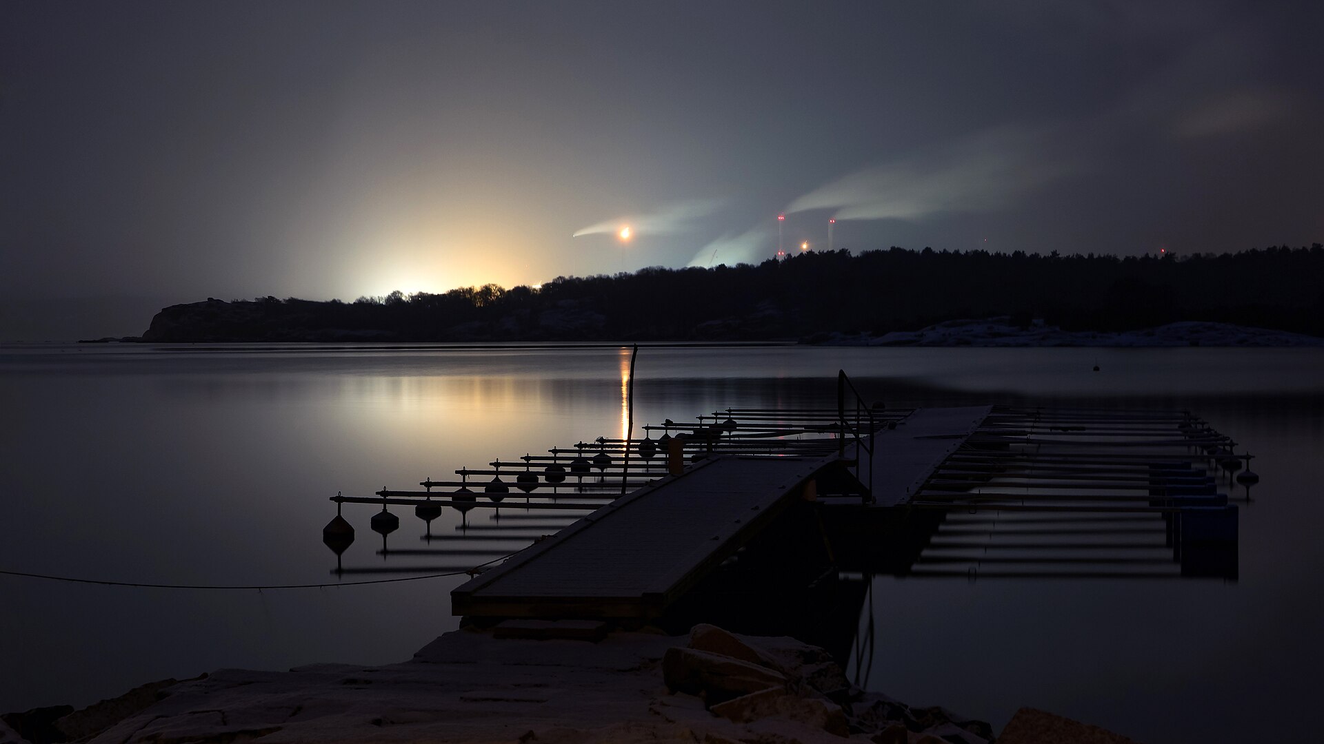 A large oil refinery seen across a fjord on a snowy winter night, its lights glowing orange and reflecting off falling snow so that the whole sky above it is lit.