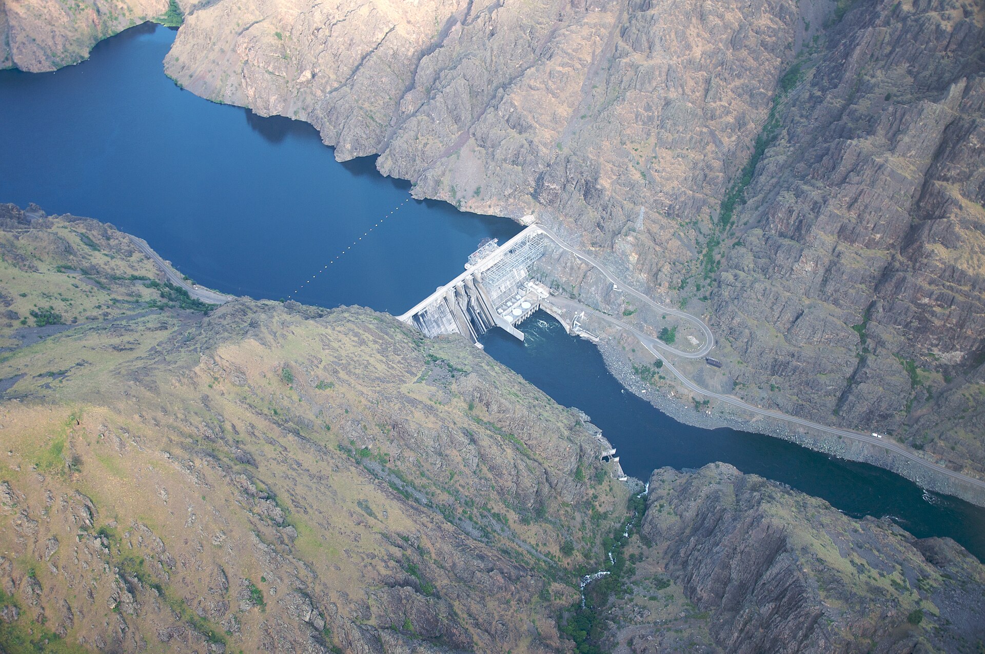 A concrete dam spanning the Snake River at the bottom of a steep, rocky, sparsely vegetated canyon, with water flowing below the dam.