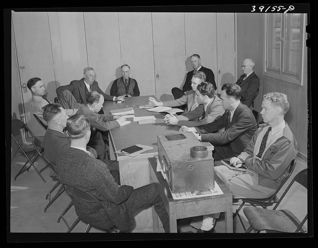 Men in suits and work clothes sit around a long table covered with papers and a metal file box, several of them talking