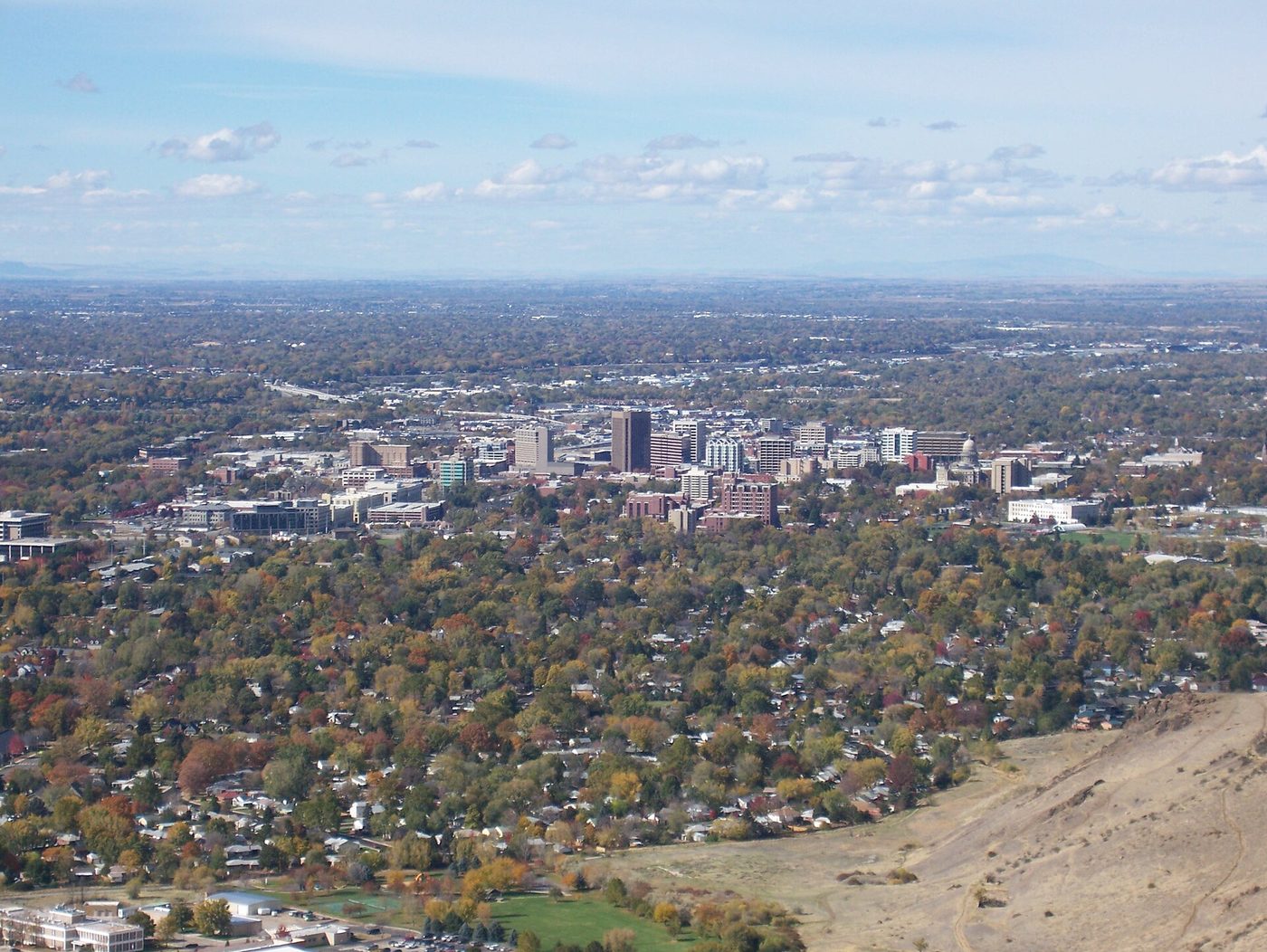 Downtown Boise office towers rise from tree-covered neighborhoods in fall color, with the flat valley floor stretching to the horizon and a bare foothill slope in the lower right corner