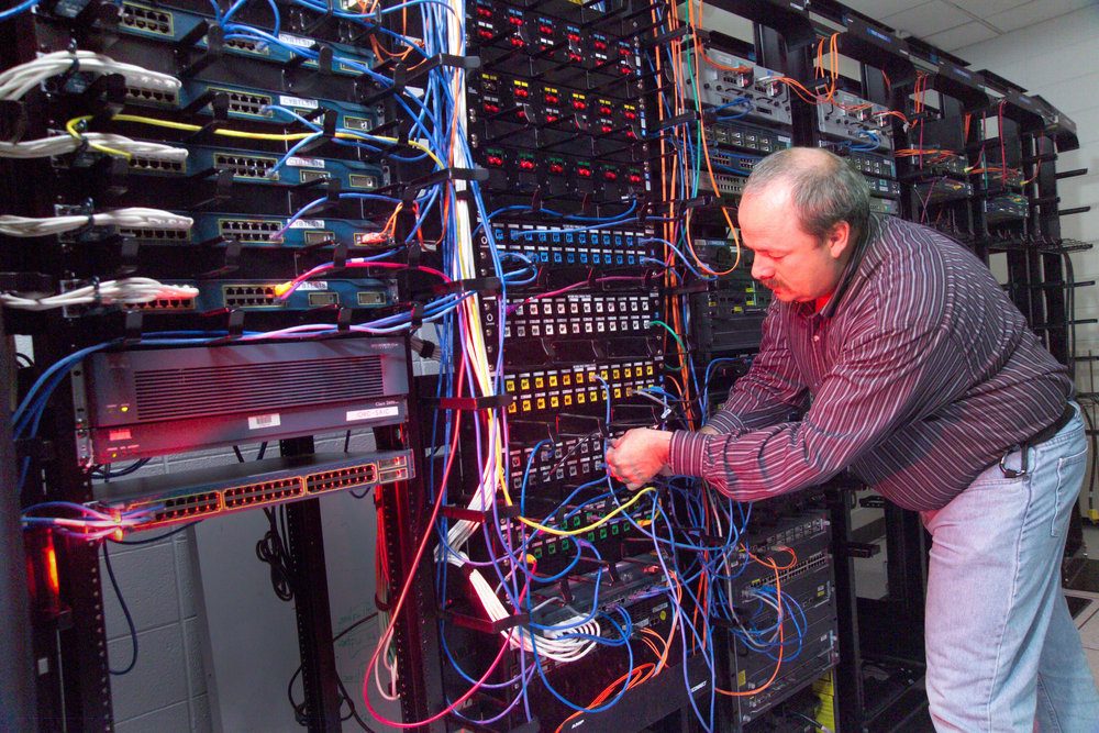 A technician connects blue and yellow network cables in a tall rack of switches and patch panels in a server room
