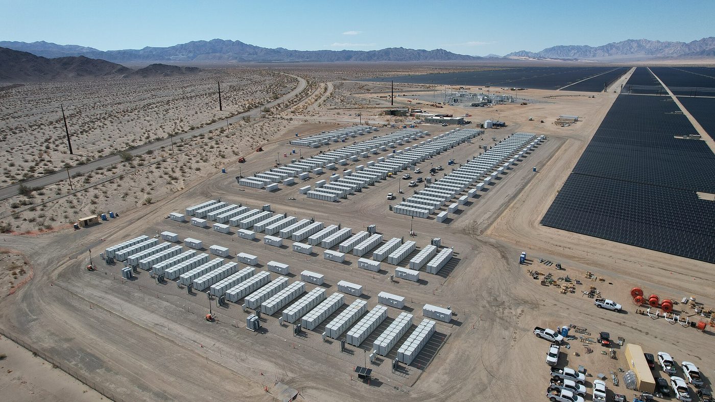 Aerial view of rows of white battery storage containers on cleared desert ground, beside a large field of dark solar panels, with mountains behind