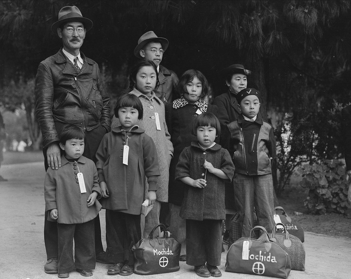 Black and white photograph of children of the Mochida family seated with identification tags attached to their coats while waiting for an evacuation bus
