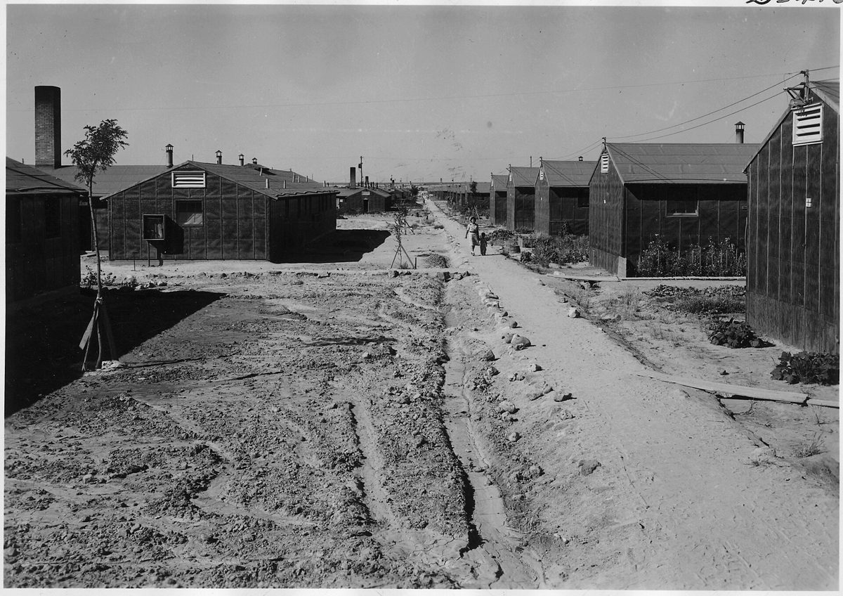 Black and white photograph looking down a dirt street between rows of tar-paper barracks at Minidoka, with small vegetable gardens beside the buildings and two people walking in the distance