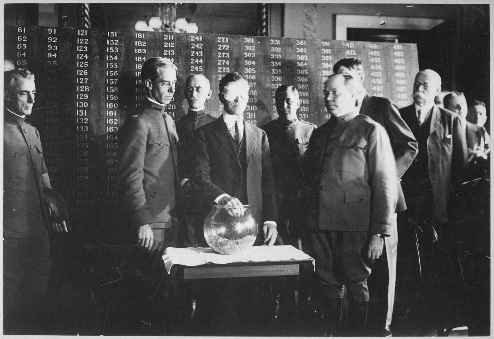 Secretary of War Newton Baker, blindfolded, drawing a capsule from a glass bowl at a draft lottery while officials watch