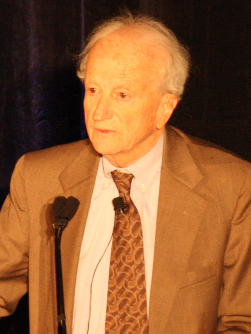 Gary Becker, an older man in a suit and glasses, speaking at a lectern