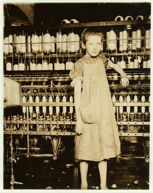 A young girl in a work dress stands barefoot beside a row of spinning frames in a cotton mill, one hand on the machine, looking at the camera.