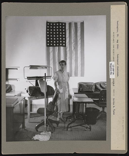 A Black woman in a work dress stands facing the camera holding a broom in one hand and a mop beside her, in front of a large American flag hanging on the wall.