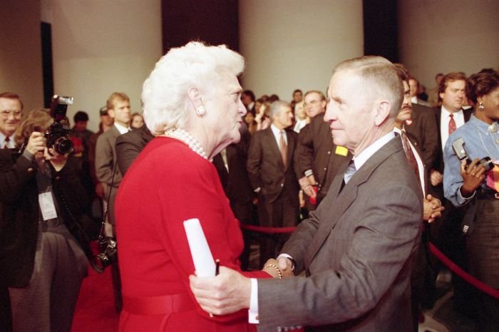 First Lady Barbara Bush, with white hair and pearls, smiling and shaking hands with Ross Perot after a televised debate