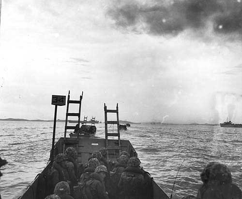 Black and white photograph of United States Marines crowded into a landing craft, some holding tall scaling ladders, heading toward a sea wall.