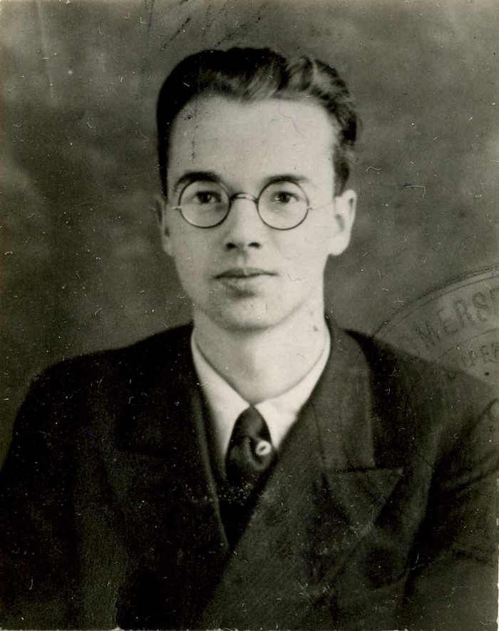 Black and white police identification photograph of a man with dark receding hair and round glasses, wearing a jacket and tie, facing the camera.