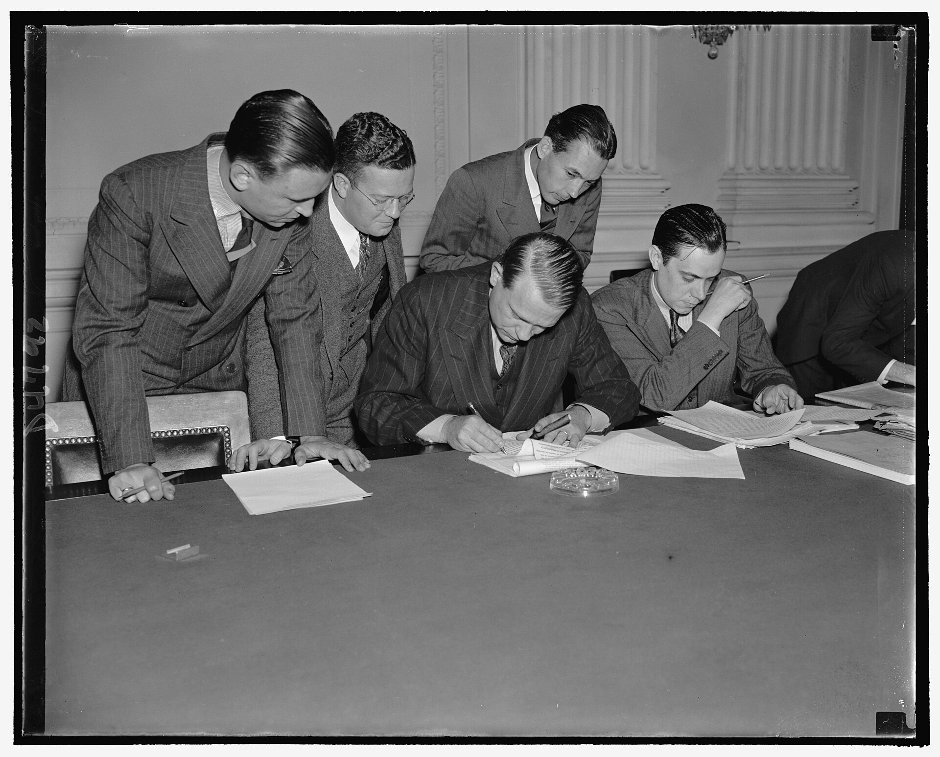 Black-and-white photograph of Martin Dies seated at a long table, pen in hand over a typed statement, while four young men in suits lean in and watch, one taking notes