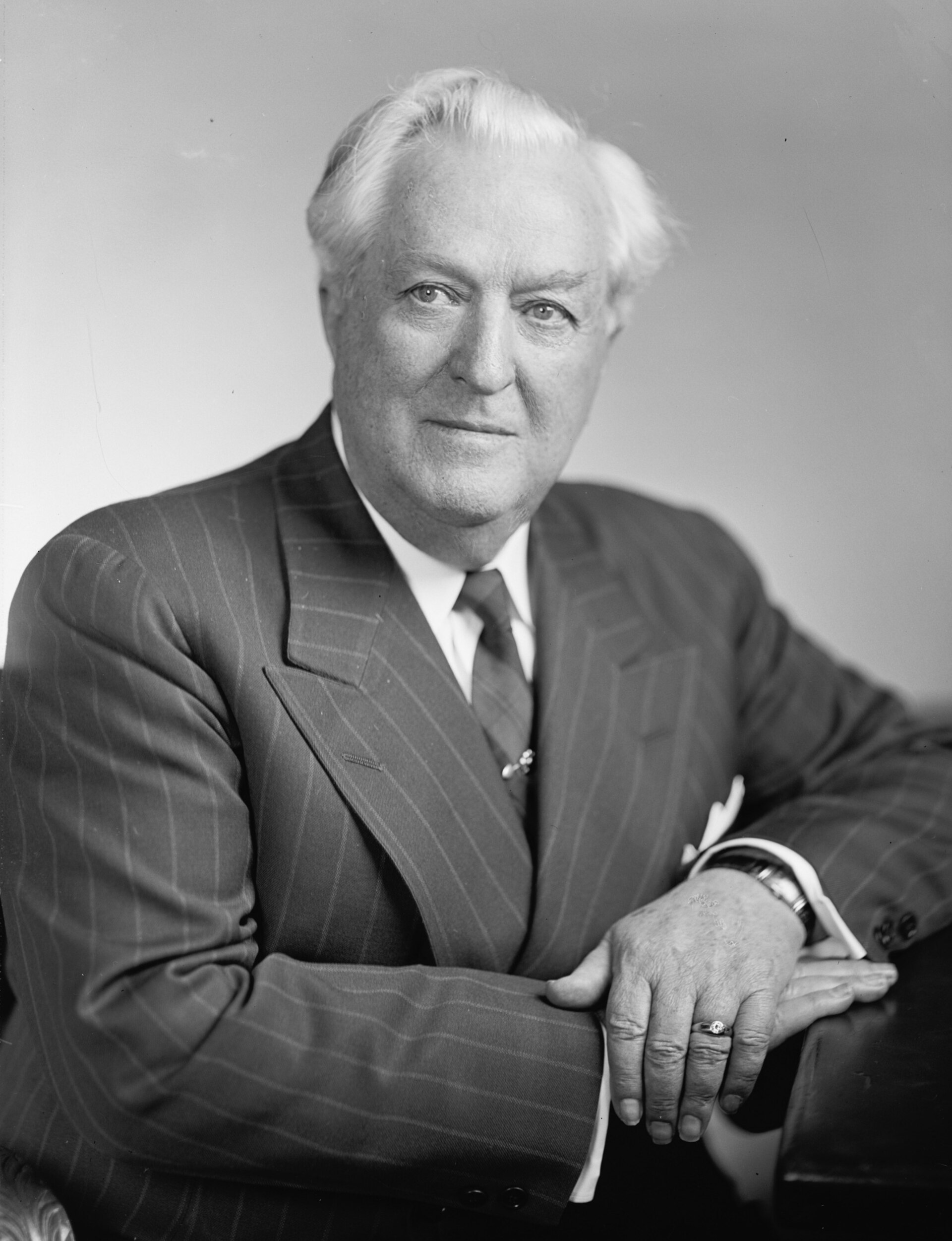 Black-and-white studio portrait of Senator Pat McCarran, a white-haired man in a pinstriped suit, seated with his hands folded on a desk