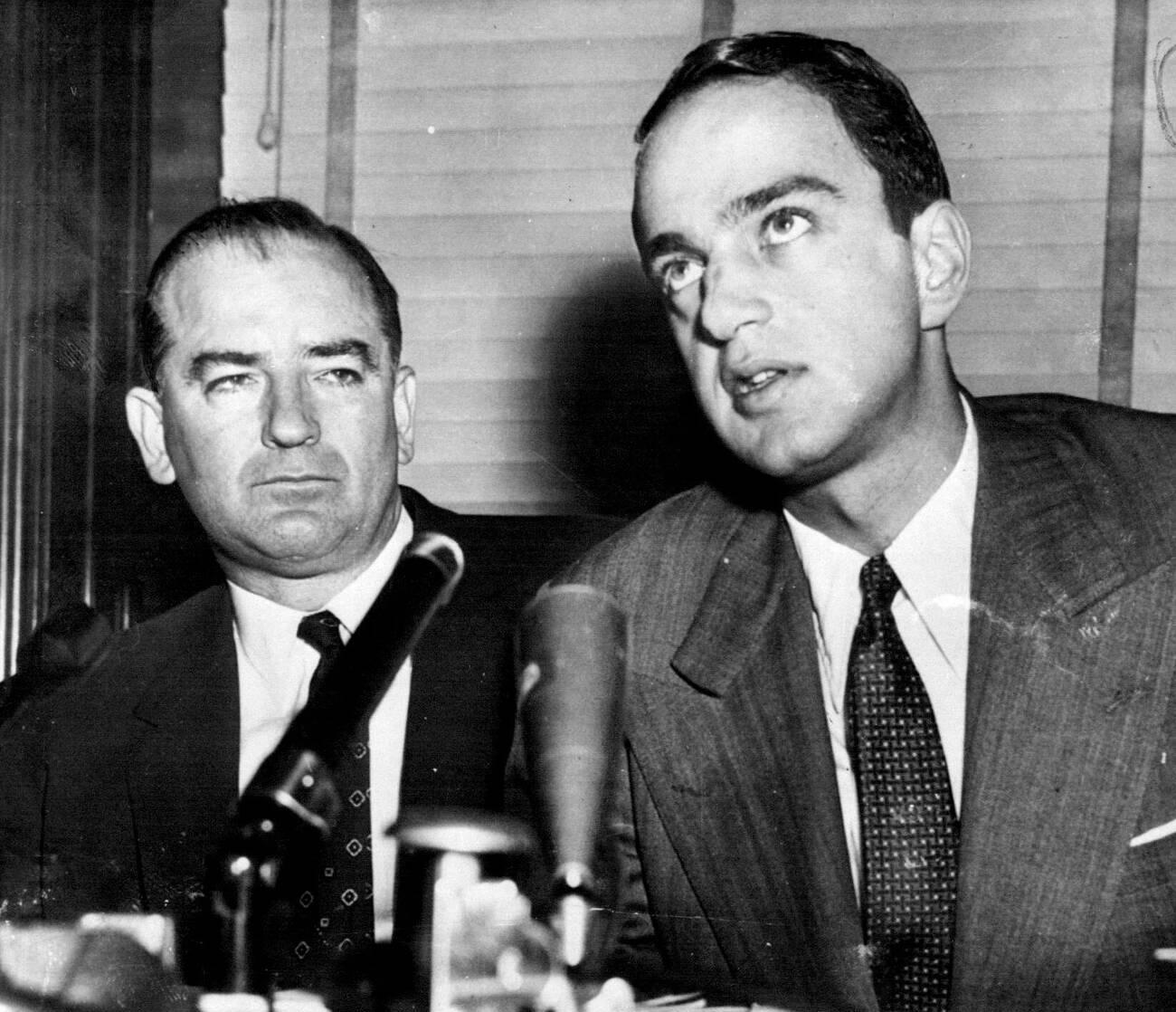 Black-and-white wire photograph of Senator Joseph McCarthy and Roy Cohn seated side by side behind microphones, Cohn speaking and McCarthy looking on