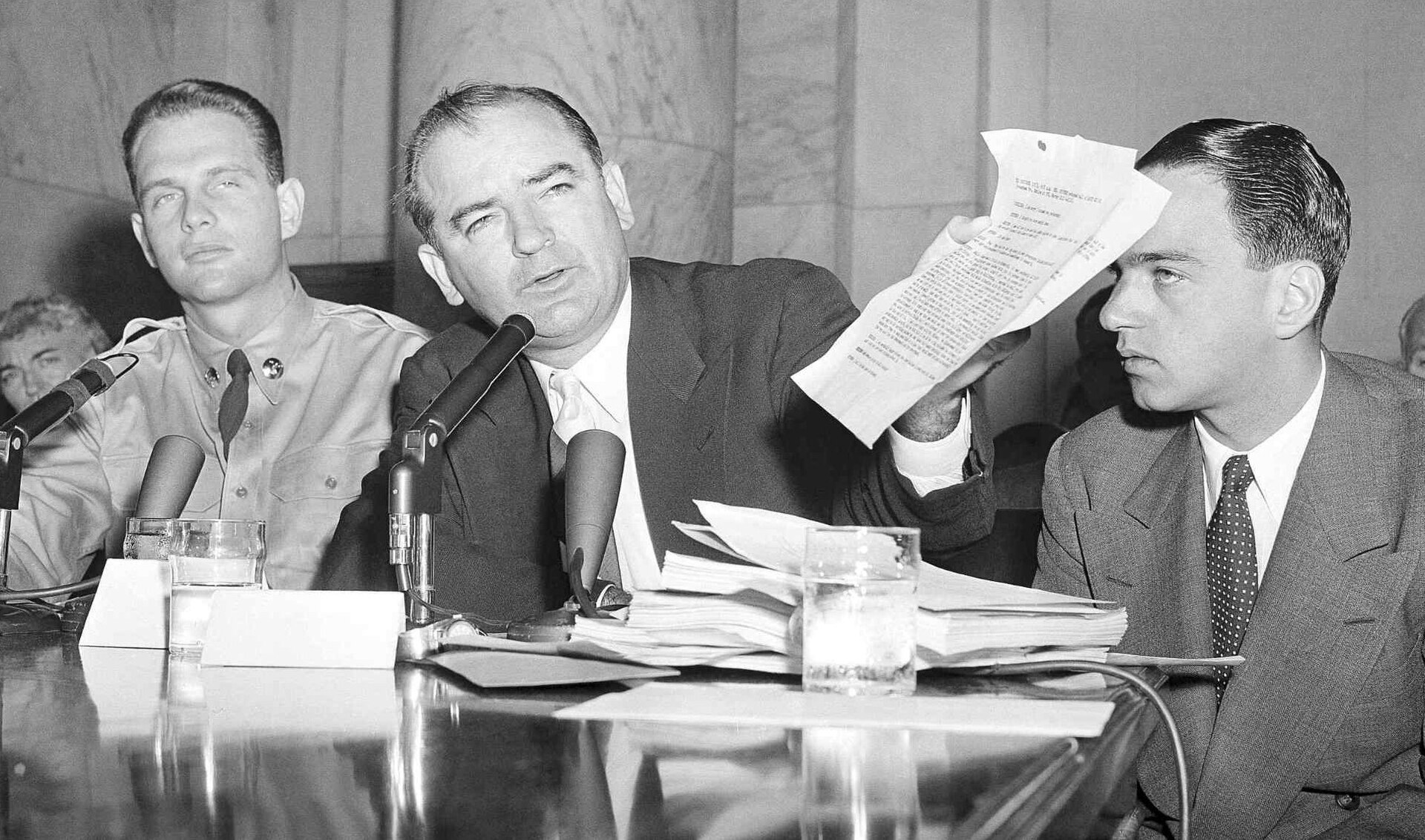 Black and white photograph of three men at a hearing table with microphones: a young man in an Army uniform at left, a man in a dark suit at center holding up a sheet of paper, and a younger man in a suit and polka-dot tie at right looking at him