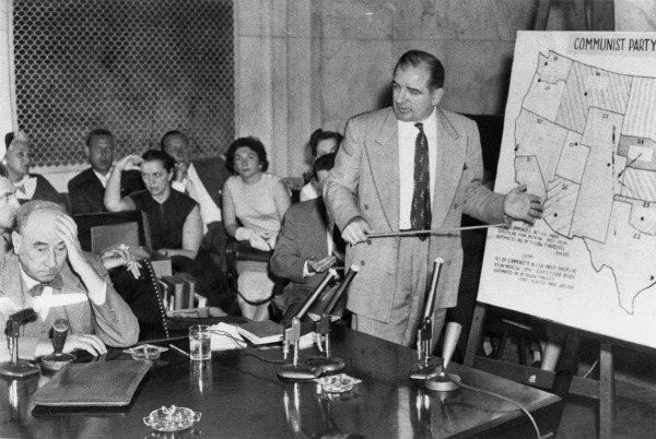 Black and white photograph of a hearing room: an older man in glasses sits at a table at left with his head resting on his hand, while a man in a light suit stands at right pointing to a large map of the United States labeled Communist Party, with spectators seated behind