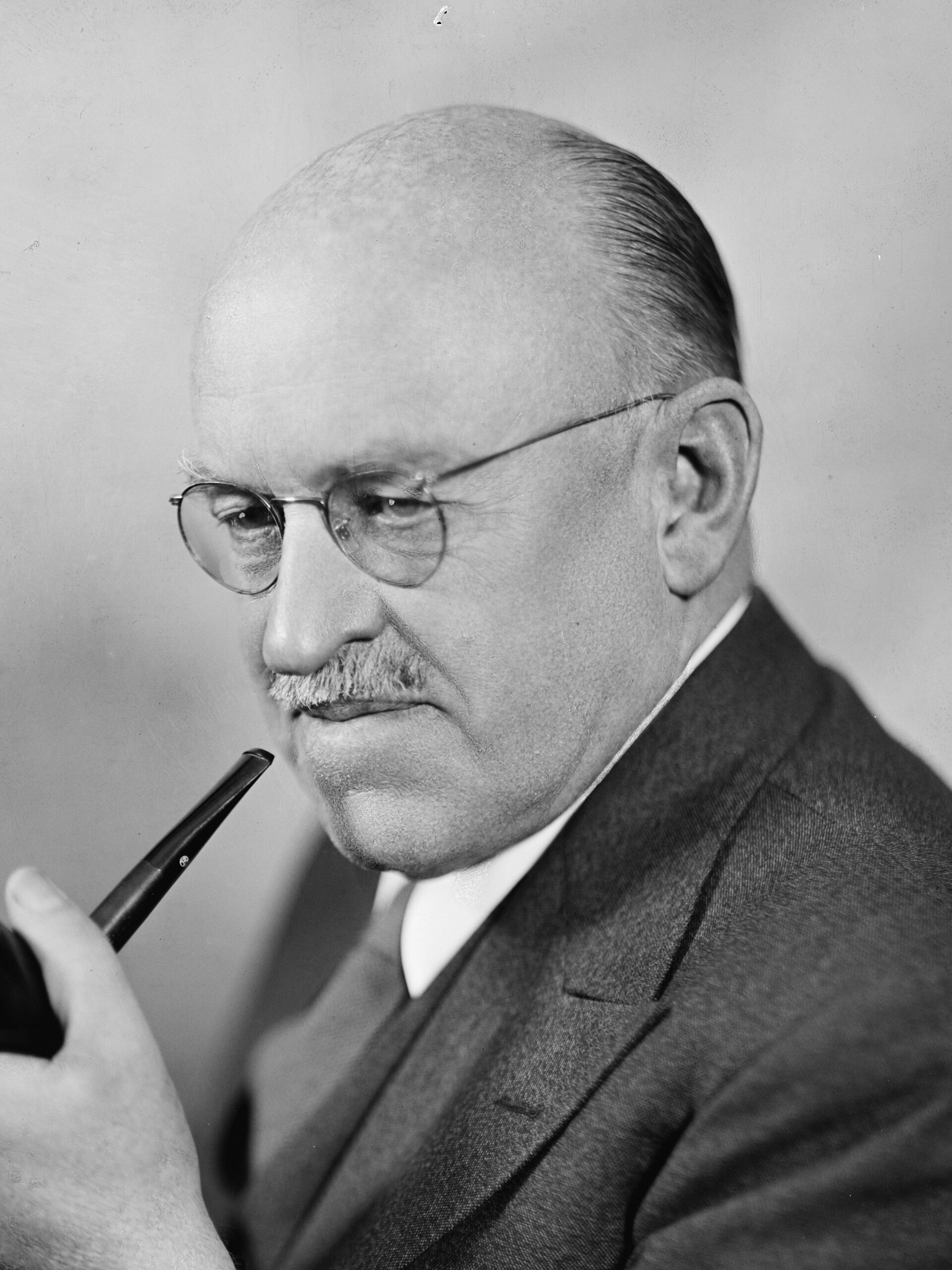 Black and white studio portrait of Ralph Flanders, a balding man with glasses and a small mustache, wearing a dark suit and holding a pipe near his chin